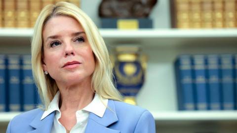 Pam Bondi looks into the distance wearing a light blue blazer and white shirt. In the background there is a white book shelf with blue and yellow books on it.