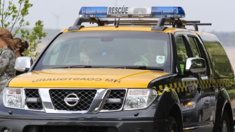 A black and yellow HM Coastguard pick-up truck with blue and white lights on the roof. Two people are sat inside. One is wearing a high-vis coat and the other has their right hand on the top of the steering wheel. Two women are stood next to the left of the vehicle.