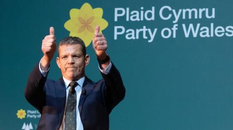 Rhun ap Iorwerth has a blue jacket over a light blue shirt. He is stood in front of a board that says Plaid Cymru in white font and Party of Wales. There is a daffodil to the right of it. He has his thumbs up. 