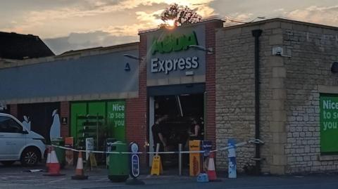An Asda express store in fading light - a police cordon around the front entrance, which no longer has it's glass or doors.