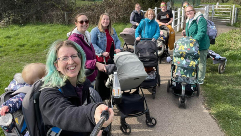 A mixed group of women with prams stand together on a paved path, smiling.