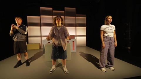 Three young actors stand on a stage looking towards the audience