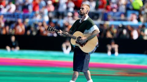Justin Bieber carrying and playing an acoustic guitar walks across a large stadium field during a live performance. He wears a dark green short-sleeved top layered over a white shirt, knee-length dark shorts, white socks. A headset microphone and in-ear monitors are visible, and tattoos can be seen on both arms and one lower leg. The field is brightly coloured with turquoise and pink sections, while a large, out-of-focus crowd fills the stadium seating in the background. The event is the FIFA World Cup 2026.