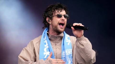 Tom Grennan is wearing dark glasses and singing into a microphone. He is wearing a light beige jacket and Coventry City scarf around his neck