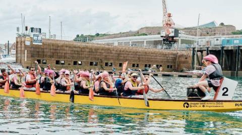 A photo that shows a number of people in a long yellow boat, with one man at the front of the boat facing the other people. He is wearing a pink hat. Some of the other people are also wearing pink hats or wigs. They are all holding oars that are out of the water.