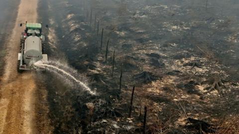 A drone view shows a farmer using his tractor to spray burnt fields and forests during a wildfire burning in the High Fens