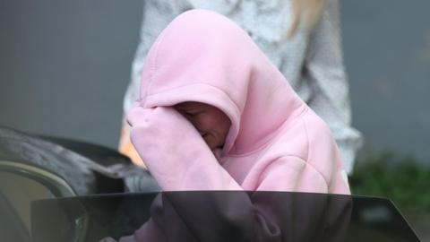 Isobella Perrie Sullivan, a woman wearing a pink hooded jacket, gets into a car following a court appearance. The hood is pulled up over her head and she is hiding her face behind her sleeve.