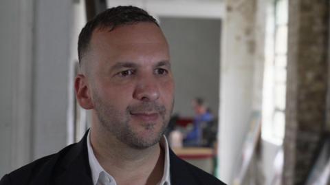 Zack Polanski being interviewed for TV. He is wearing a dark jacket and a white open-necked shirt. There is an blurred background image with a window to one side. 