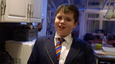 A year 7 student wearing uniform and tie, smiling, in the kitchen at their house.