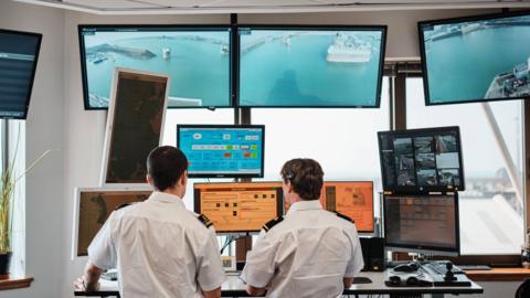 Two men in white short sleeved maritime shirts stand in a control centre looking at screens of video footage and information of the Jersey coast.