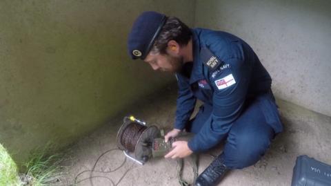 A picture of a Royal Navy officer crouched down holding a remote. He is in the corner of a room.