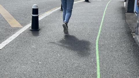 Green line running along a pavement. A pedestrian walks next to the line away from the camera - only their legs in jeans and feet in trainers can be seen - and their shadow can be seen behind them. A bollard on the left of the picture is at the edge of the pavement next to a yellow line.
