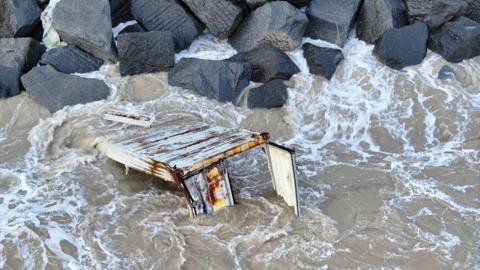 A rusty, white shipping container floating in the sea. It is extensively damaged, missing its top and bottom and with a broken door. It is mostly submerged in water, and appears stuck on a wall of large black rocks.