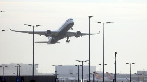 A plane takes off over Heathrow airport. Buildings can be seen in the background and other planes are parked alongside them 