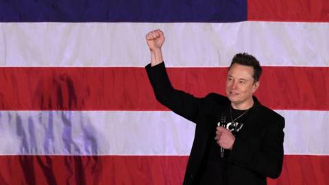 Musk standing in front of a US flag with his fist raised