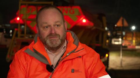 A man dressed in a bright orange jacket with the Hull City Council logo on its right-hand side. He has short brown hair, a cropped beard and moustache and blue eyes.  
