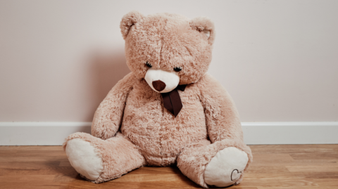 Beige teddy bear with dark brown neck-tie sits on a wooden floor