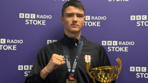 A teenage boy with short black hair, a black top and a medal around his neck is holding his right hand up in a fist while holding a gold trophy in his left hand. He is standing in front of a purple background with "BBC Radio Stoke" in white letters.