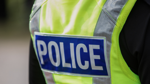Stock image of the back of a police's florescent jacket that reads POLICE. The man wearing the jacket with a black t-shirt.