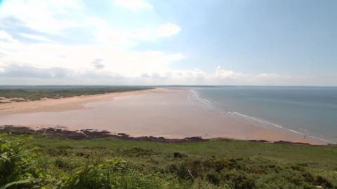 Saunton Sands at low tide is a huge sandy beach with a handful of walkers. The sea is flat and the sky sunny with clouds.