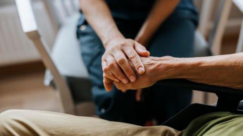 A care worker holds the hand of an elderly patient in a close-up image. The care worker's blue uniform can be seen and she is sitting on a chair.