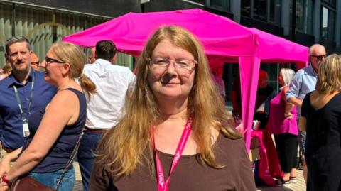 Lesley Wood with long blond hair and glasses wearing a brown t-shirt. She stands in front of a handful of protesters in Liverpool city centre. She is smiling.