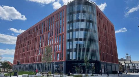 The seven-storey building with red terracotta tiles around the windows on each side and a large rounded glass corner over the entrance that stretches from the first floor to the roof.