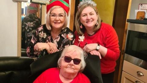 Two women - Brendya young and her sister Janice - wearing Christmas hats, behind their mother Margaret Chisholm who also has Christmas-related items on her head, and dark glasses.