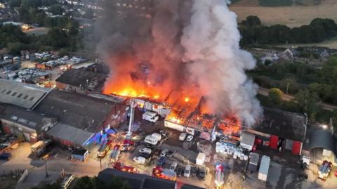 An aerial view of a huge seat of fire across a large industrial complex with a plume of smoke rising high