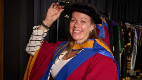 Cockroft wears a university cap and gown. The gown is red with yellow and blue trims. She is smiling and has long blonde hair.