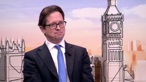 Alex Burghart, who has short brown hair and wears black-rimmed glasses, wears a blue tie with a white shirt and black suit on Laura Kuenssberg