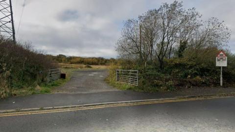 View taken from a rural road of an unmade road leading though two open six-bar gates into an area of open scrubland. Trees and shrubs can be seen on either side of the gates, and also beyond the open area.