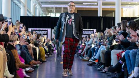 Stuart Trevor on the catwalk at one of his shows wearing red and blue check trousers, trainers and blue decorated jacket with an audience sat around him taking photos on their phones