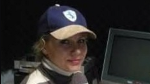 A woman with blonde hair and a blue and beige baseball cap is smiling in front of a monitor