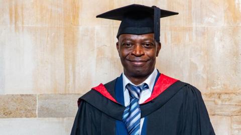 A man smiles while wearing a graduation gown and hat.
