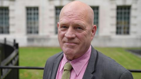 Stephen Close smiles at the camera infront of a green grassy lawn, black railings and a light-coloured-brick building in Whitehall, central London. He wears a pink shirt, sage green tie and a grey suit.