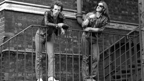 A black and white picture of Lucas Fox and Lemmy Kilmister standing on a staircase outside a building.