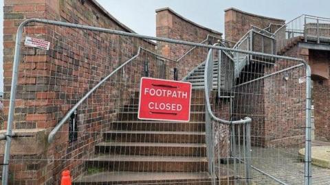 A metal fence placed in front of the steps leading up to Irish Gate Footbridge in Carlisle, with a sign indicating the footpath is closed.