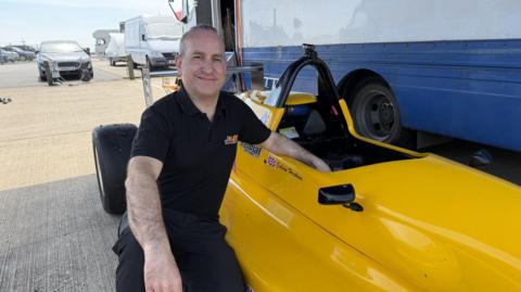A man in a black polo shirt kneels beside a bright yellow single-seat race car in a paddock area. The car has a small British flag and text reading “British Racing & Sports Car Club.” Behind them are transport trucks, parked cars and equipment.