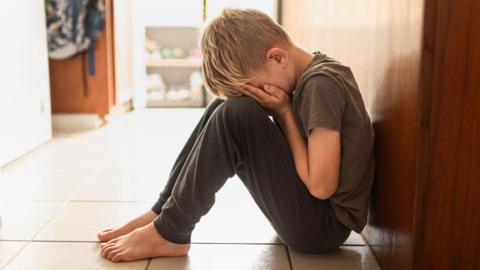 A stock image of a child sat on a tile floor covering their eyes. They have their head on their knees. It is a young boy, with short blonde hair, and he is wearing a grey t-shirt and a pair of grey tracksuit bottoms.