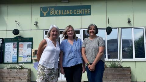 Three middle-aged women stand in front of a pale green building. A sign reading "The Buckland Centre" is on the wall behind them.