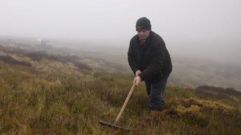 A man in a black jacket and black hat is using a rake to pull moss out of the ground, he is also wearing dark trousers. In the background there is moss covering the entire ground in various colours including green, brown and yellow, the sky is full with fog. 