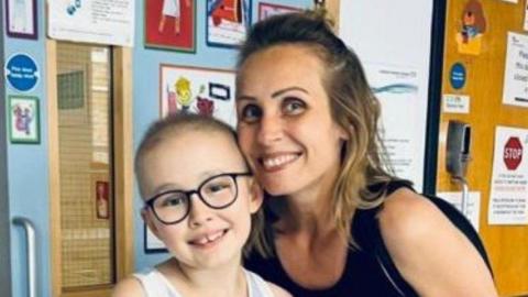 Christina and Skye. Christina is wearing a black sleeveless top and Skye a white one. They are in a hospital corridor which has been decorated with posters.