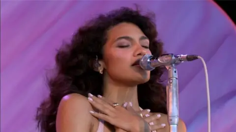 A close-up of Olivia Dean. She has her eyes closed and is singing into a silver microphone. She has her hands crossed and they are resting just below her neck and she has pale pink nail varnish on and a gold ring on one finger. The backdrop is purple