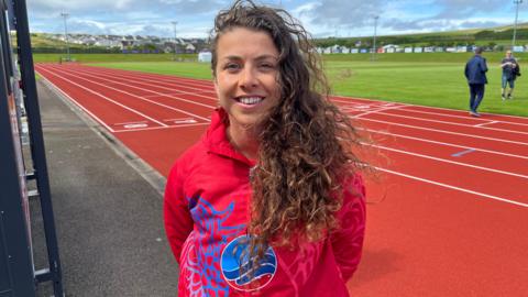 A woman with long, curly brown hair swept to one side stands in front of a red athletics track. She is wearing a red tracksuit top.