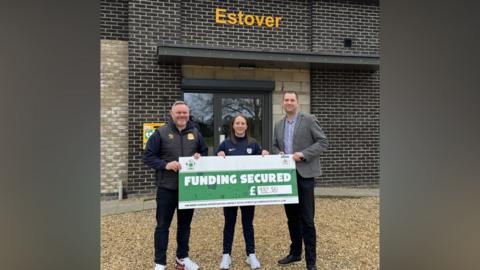 Two men and a woman smile at the camera as they hold a large cardboard cheque that reads: "Funding Secured £982,381". They are standing on gravel and in front of a building.