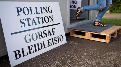An image of a polling station viewed from below, with a large sign that says "polling station" in Welsh and English. A person wearing jeans can be seen walking out of the polling station door to the right of the picture.
