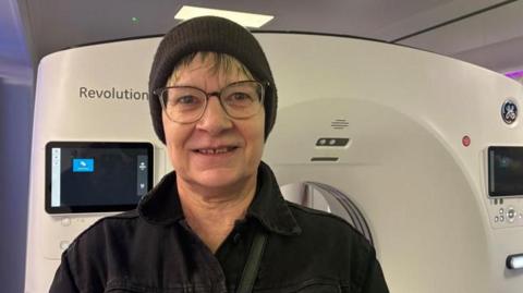 Tracey Markham stood in front of the CT scanner inside the mobile unit. She has a black beanie-style hat on and wears a black jacket.