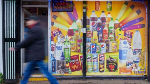 A man walks in front of a convenience store window with product advertising posters on it