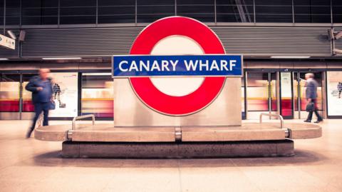 A Tube station sign saying Canary Wharf on the platform in an Underground station. A train is arriving at the platform behind. 